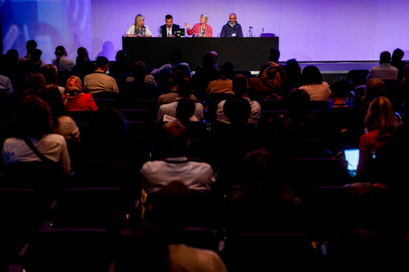 At a bustling London conference, a panel of four speakers sits at a long table on stage against a vivid purple backdrop. The audience listens intently, with several attendees taking notes as the conference photographer captures these engaging moments. James Gifford-Mead Photography - Event Photographer London