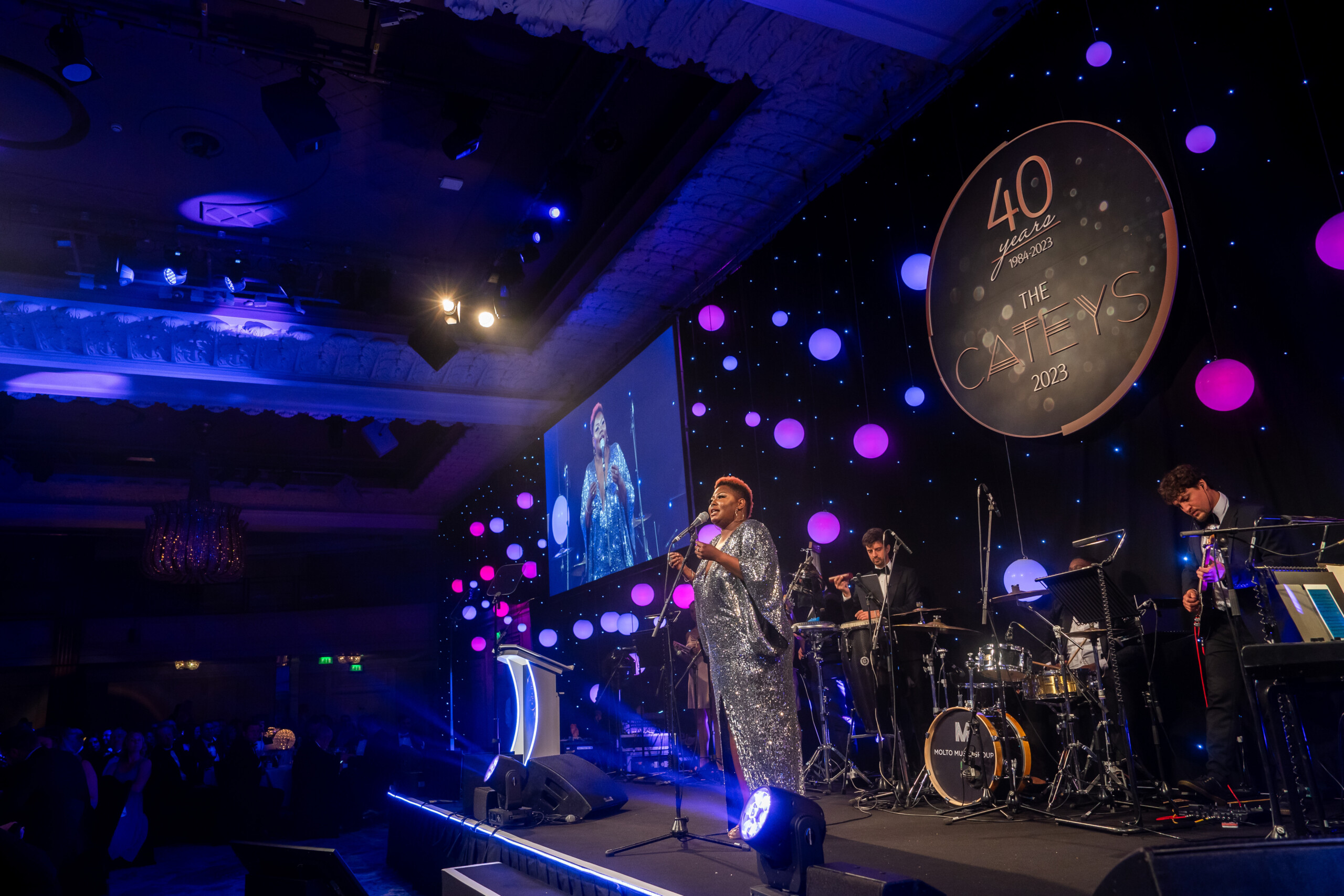 A performer in a sparkly outfit sings on stage at a gala event, where an awards photographer captures the moment. The stage is adorned with round lights and a sign reading "40 Awards, The CA News 2023," complemented by a band and a screen displaying the vibrant scene. James Gifford-Mead Photography - Event Photographer London