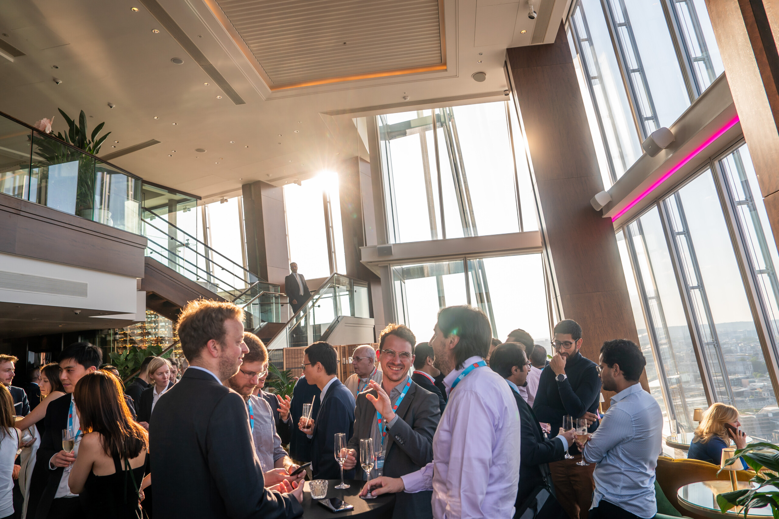 People gathered at a modern rooftop event with tall windows and a city view. They are socializing and holding drinks, while sunlight streams in through the glass. The atmosphere is lively and upscale. James Gifford-Mead Photography - Event Photographer London