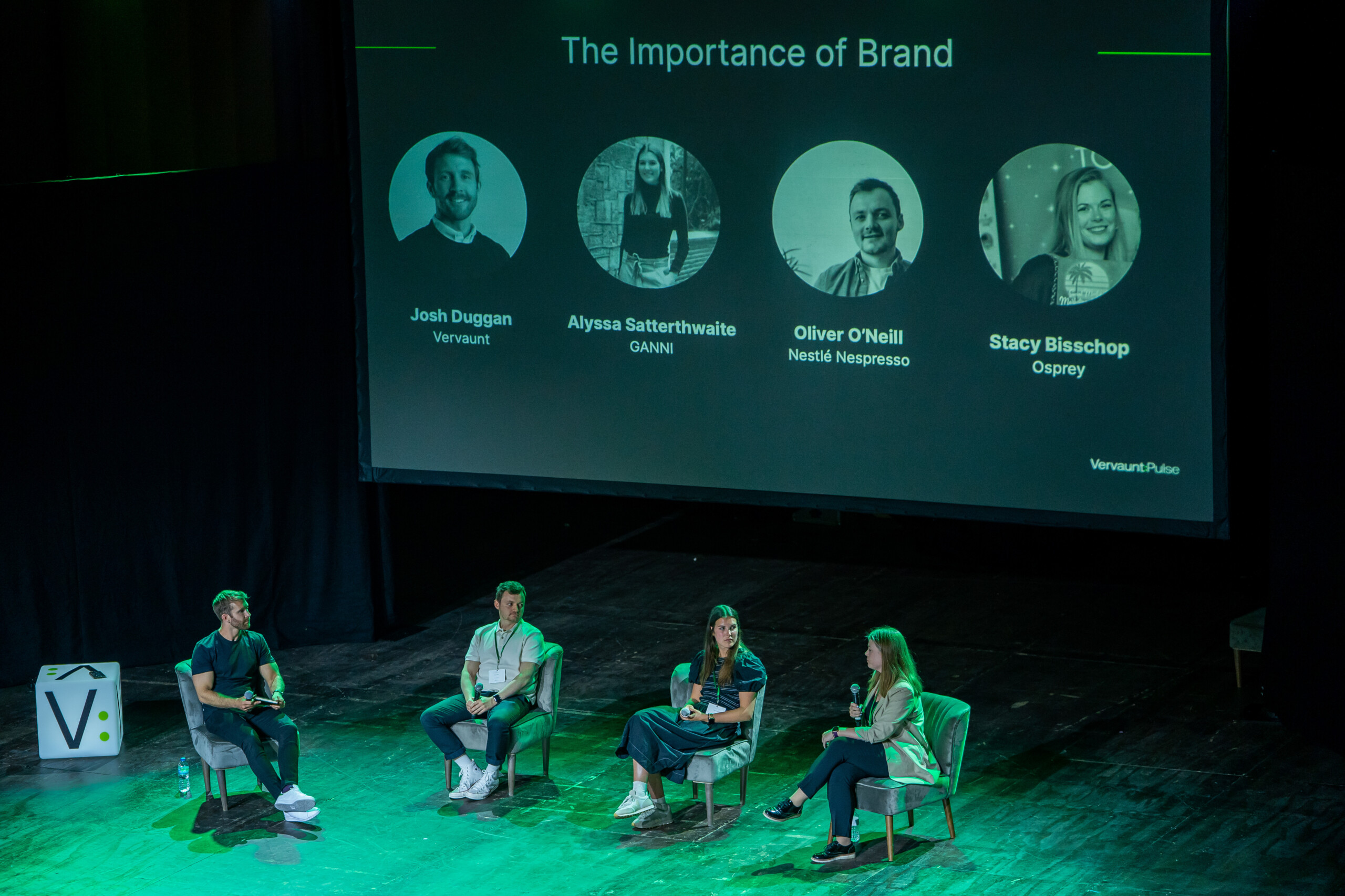Four people are seated on a stage in front of a large screen labeled "The Importance of Brand," showing four headshots and names. The setting appears to be a panel discussion or presentation. James Gifford-Mead Photography - Event Photographer London