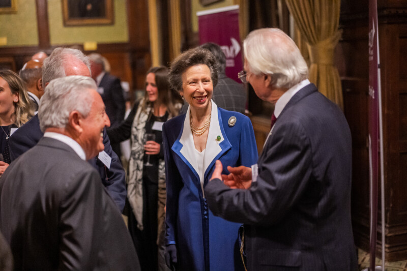 A woman in a blue coat smiles and converses with several people in a formal setting, captured by a London conference photographer. The room's wooden paneling and portraits set the scene, while other guests gather to add to the lively atmosphere. James Gifford-Mead Photography - Event Photographer London