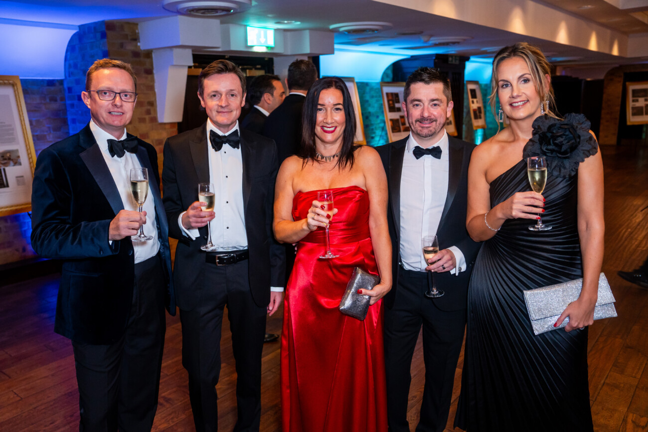 A group of five people, dressed in formal evening wear, are standing indoors holding glasses of champagne. The two women are wearing elegant gowns, and the three men are in tuxedos. The setting appears to be a formal event or gathering. James Gifford-Mead Photography - Event Photographer London