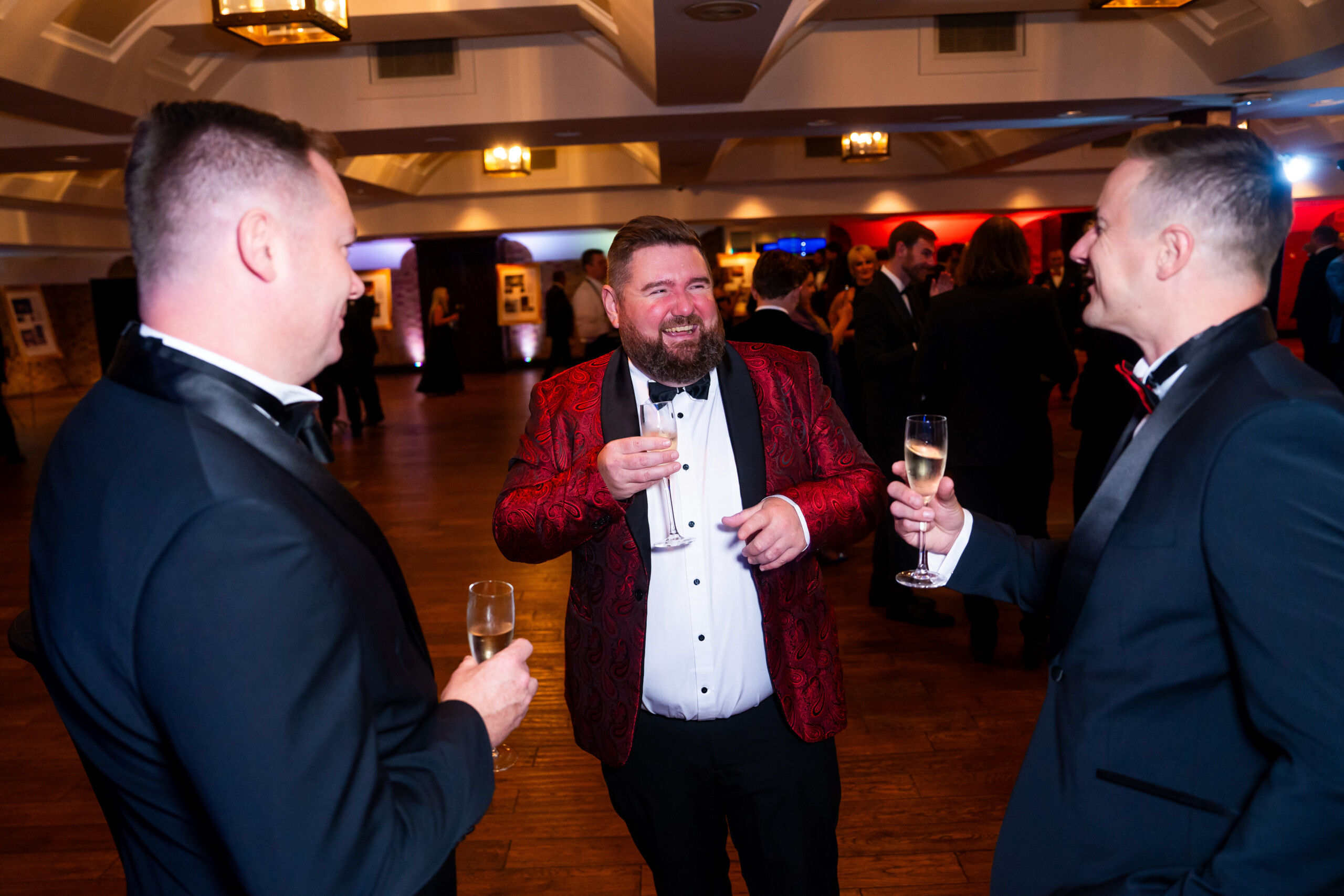 Three men in formal wear enjoy drinks and conversation at an elegant indoor event. One man wears a striking red patterned jacket, while the others wear black tuxedos. The warmly lit room is bustling with people in the background. James Gifford-Mead Photography - Event Photographer London