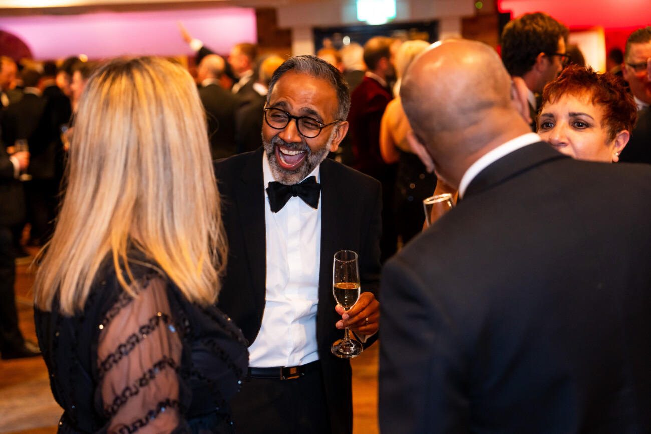 A group of people in formal attire are at a lively social event. A person in a tuxedo and bow tie is laughing while holding a champagne glass. Others around them are engaged in conversation, with blurred guests in the background. James Gifford-Mead Photography - Event Photographer London