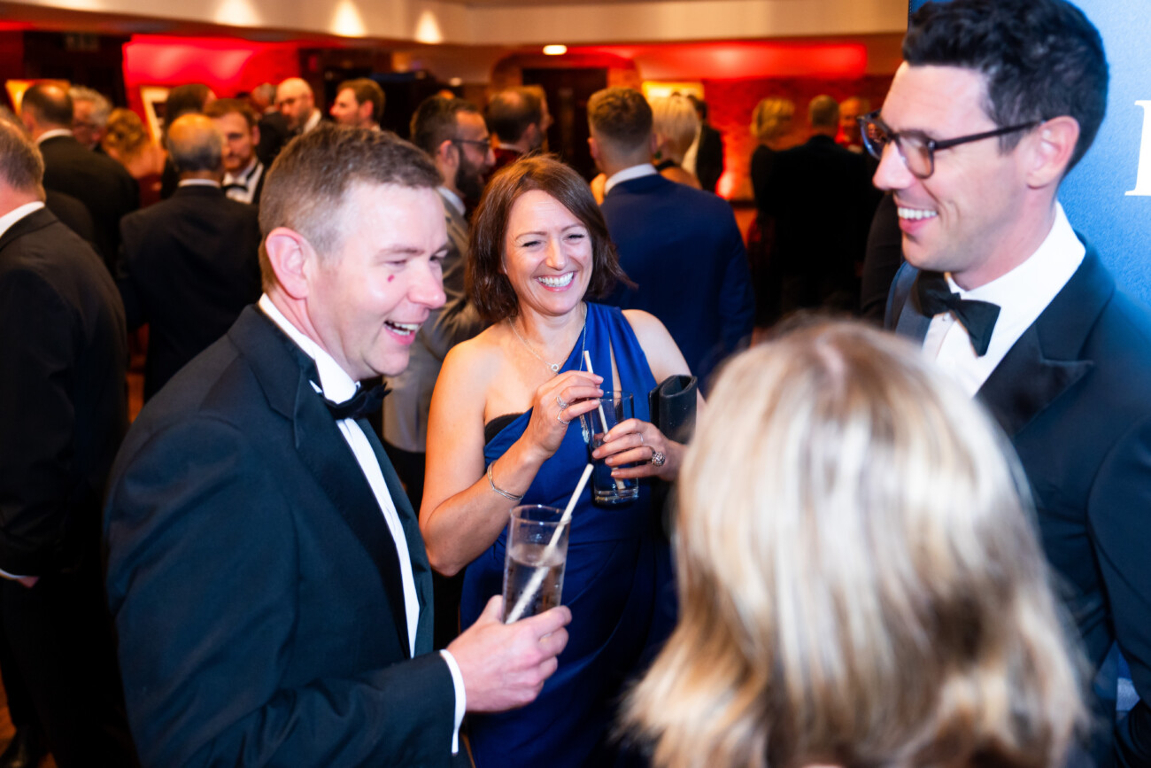 A group of people in formal attire enjoying a lively conversation at a social event. A man and a woman are holding drinks and smiling, while another man, wearing glasses, is laughing. The background is bustling with other guests. James Gifford-Mead Photography - Event Photographer London