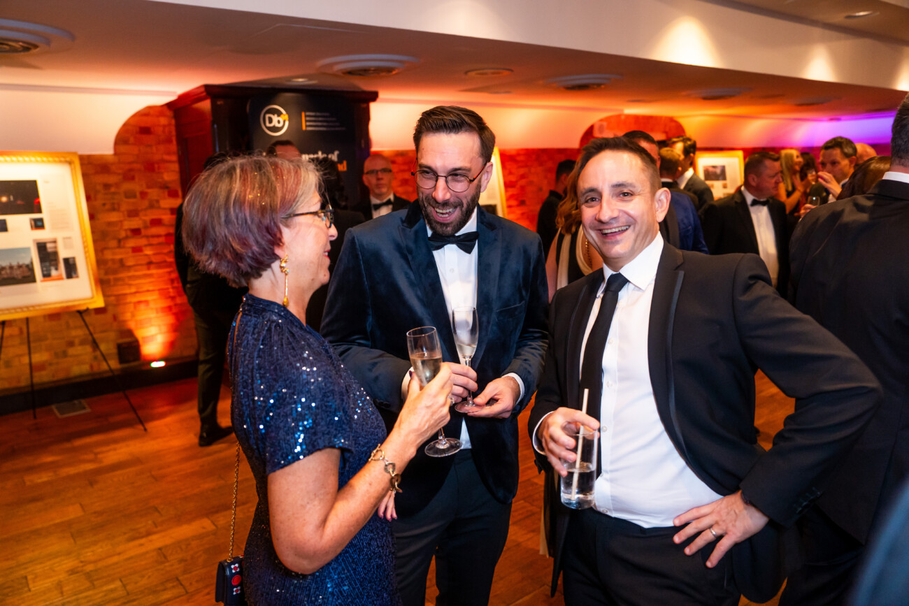 Three people in formal attire are smiling and conversing at an indoor event. One person is holding a champagne glass. The background features a framed picture, brick walls, and warm lighting. James Gifford-Mead Photography - Event Photographer London