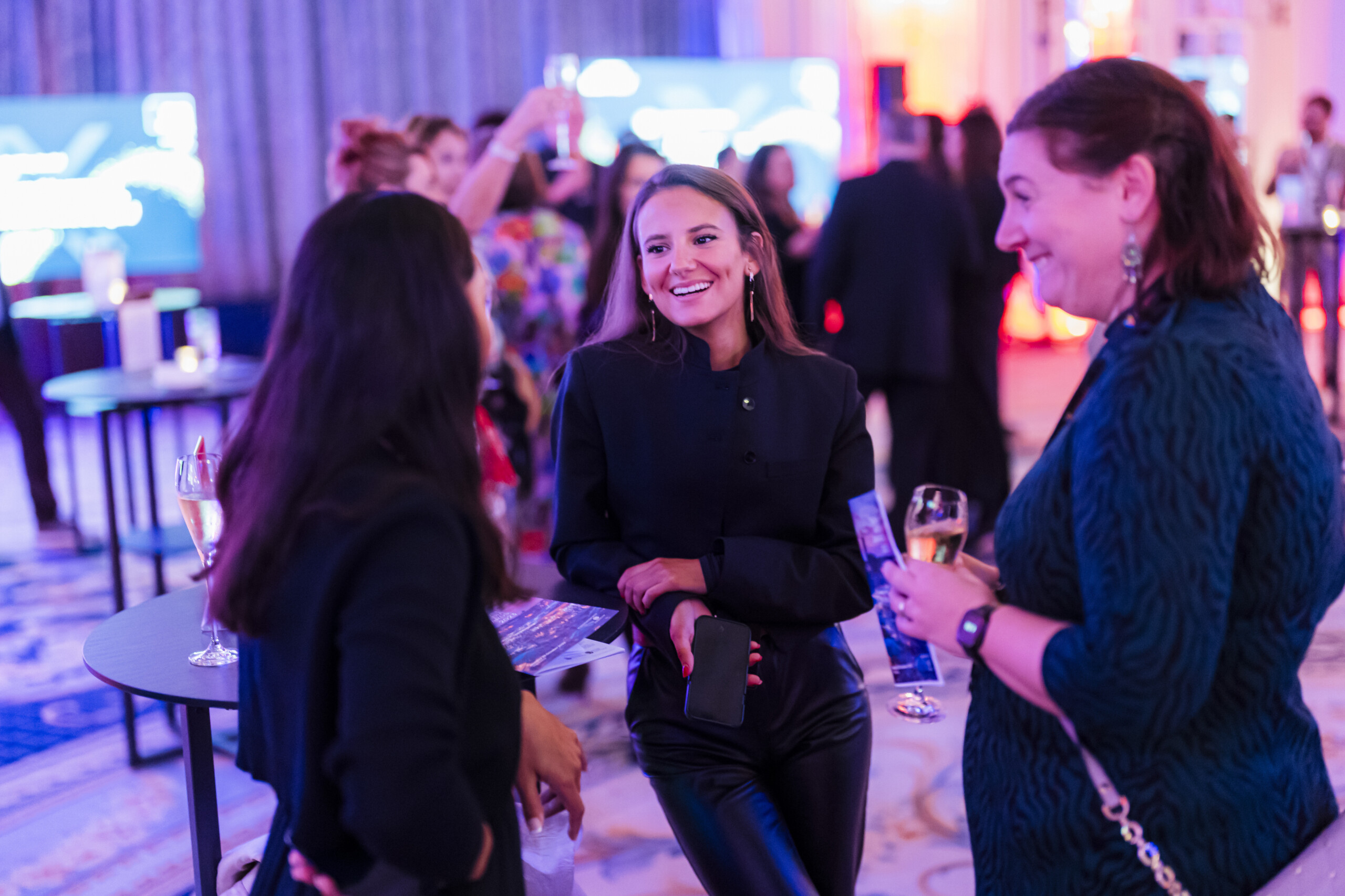 Three women in conversation at a formal event. They are holding glasses and smiling, dressed in elegant attire. The setting is a brightly lit venue with other attendees and tables in the background. James Gifford-Mead Photography - Event Photographer London