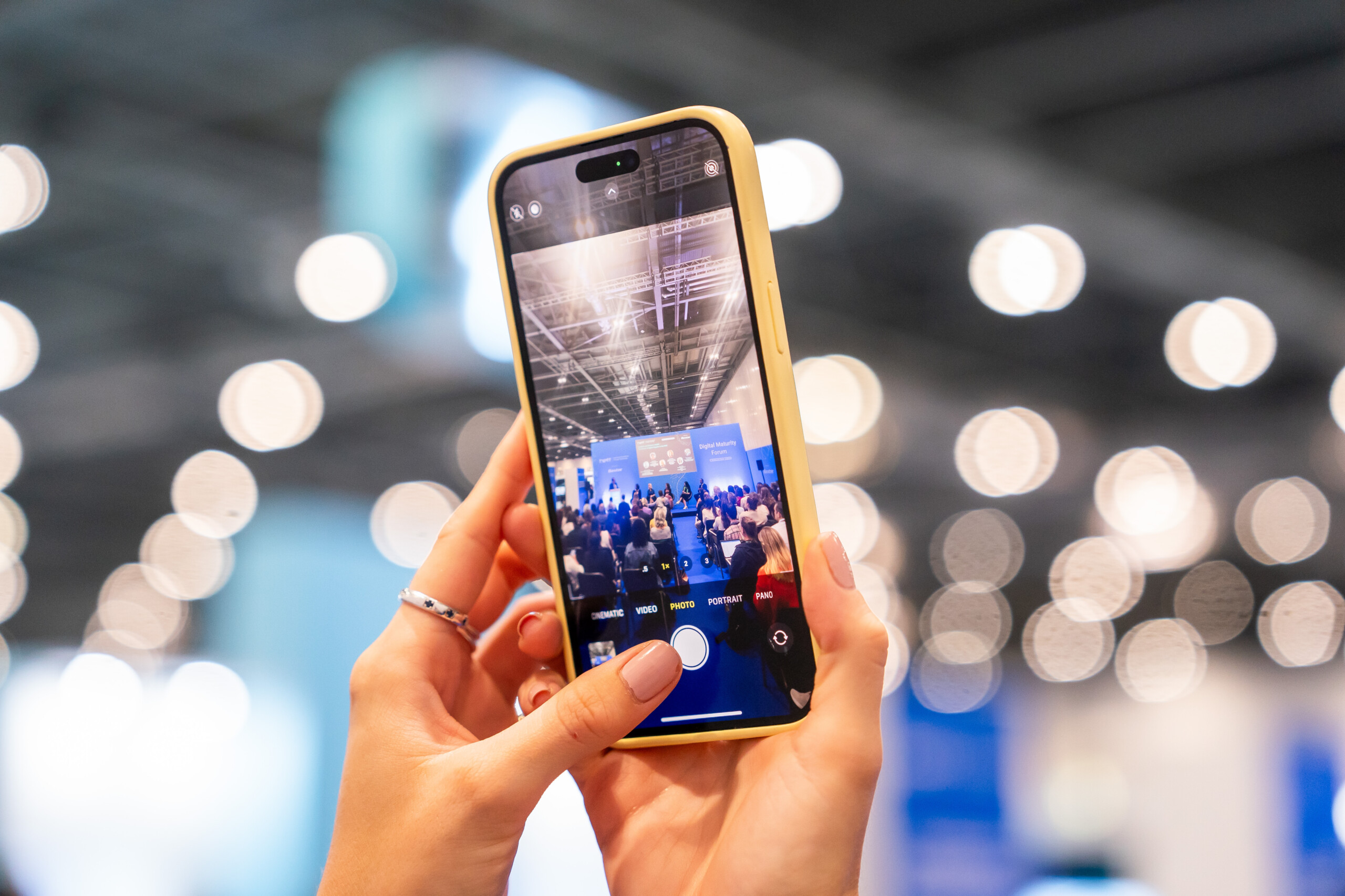 A person is holding a smartphone with a yellow case, capturing a video or photo of a conference or event. The background is blurred with bokeh lights and an audience visible on the phone screen. James Gifford-Mead Photography - Event Photographer London