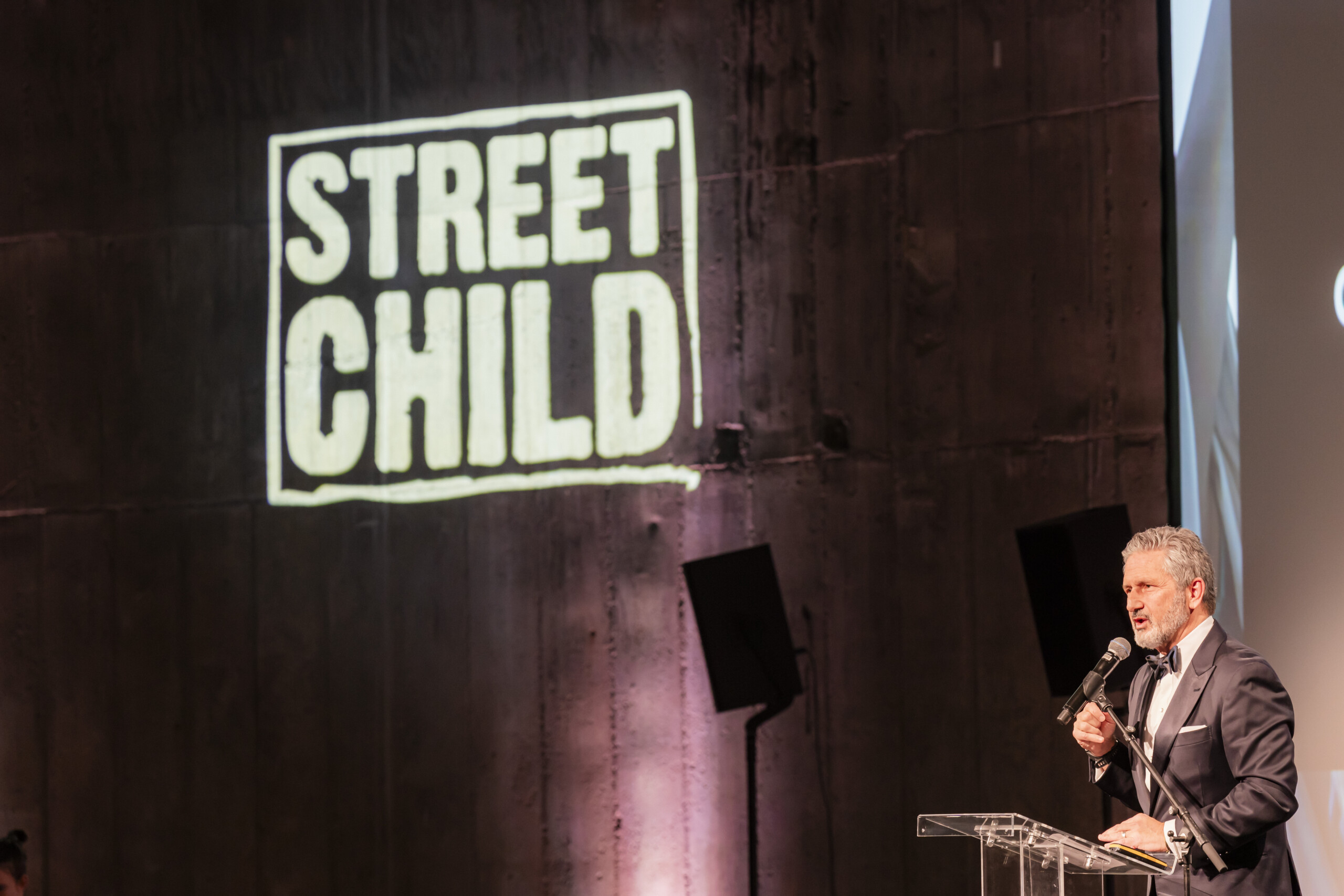 A man in a suit speaks at a podium in front of a textured wall with the illuminated logo "Street Child." The setting appears to be a formal event or conference. James Gifford-Mead Photography - Event Photographer London