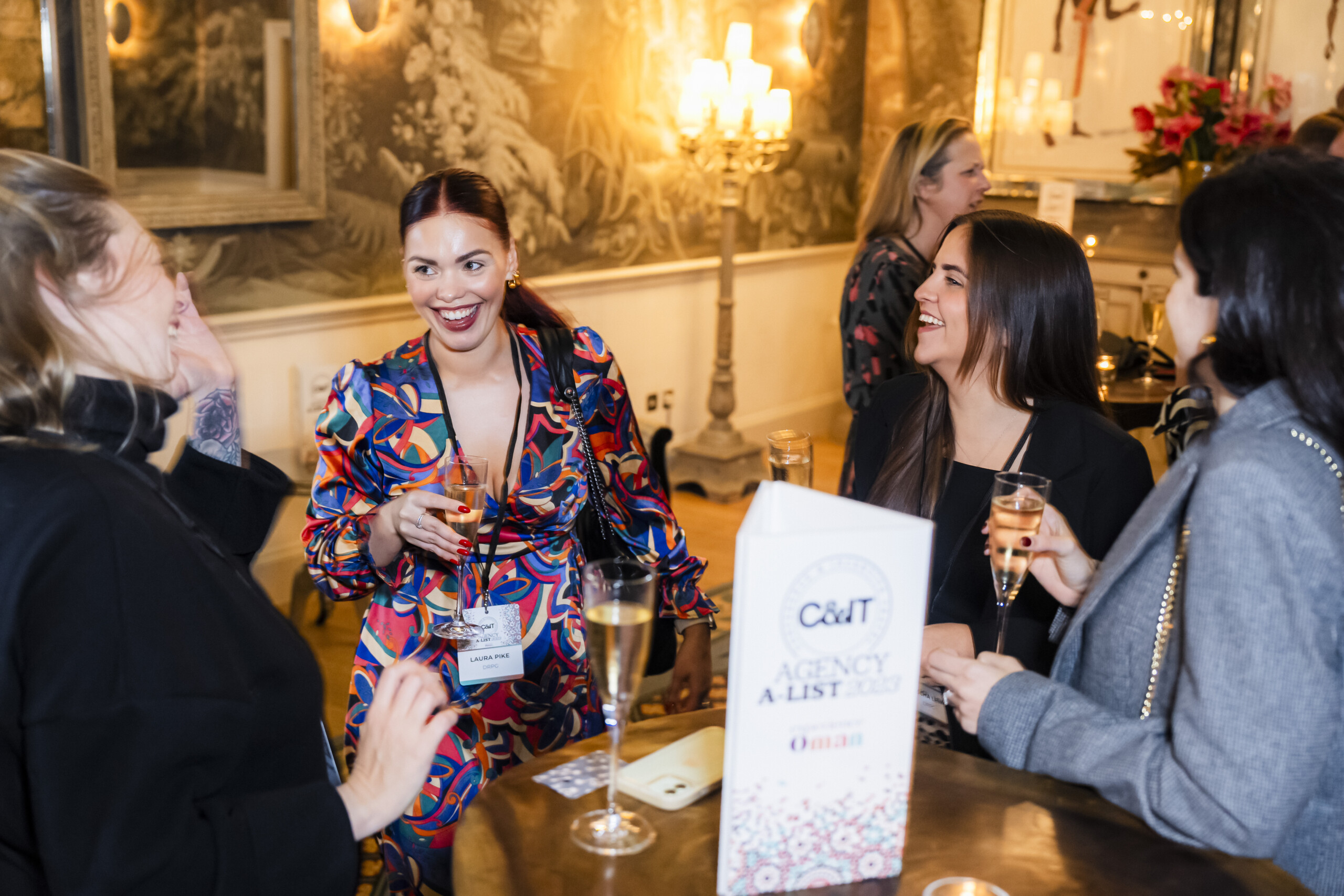 A group of four people stand around a table, smiling and chatting at an indoor event. One person holds a glass of champagne. A sign on the table reads "C&IT Agency A-List 2023." The background features decorative wall art and a chandelier. James Gifford-Mead Photography - Event Photographer London