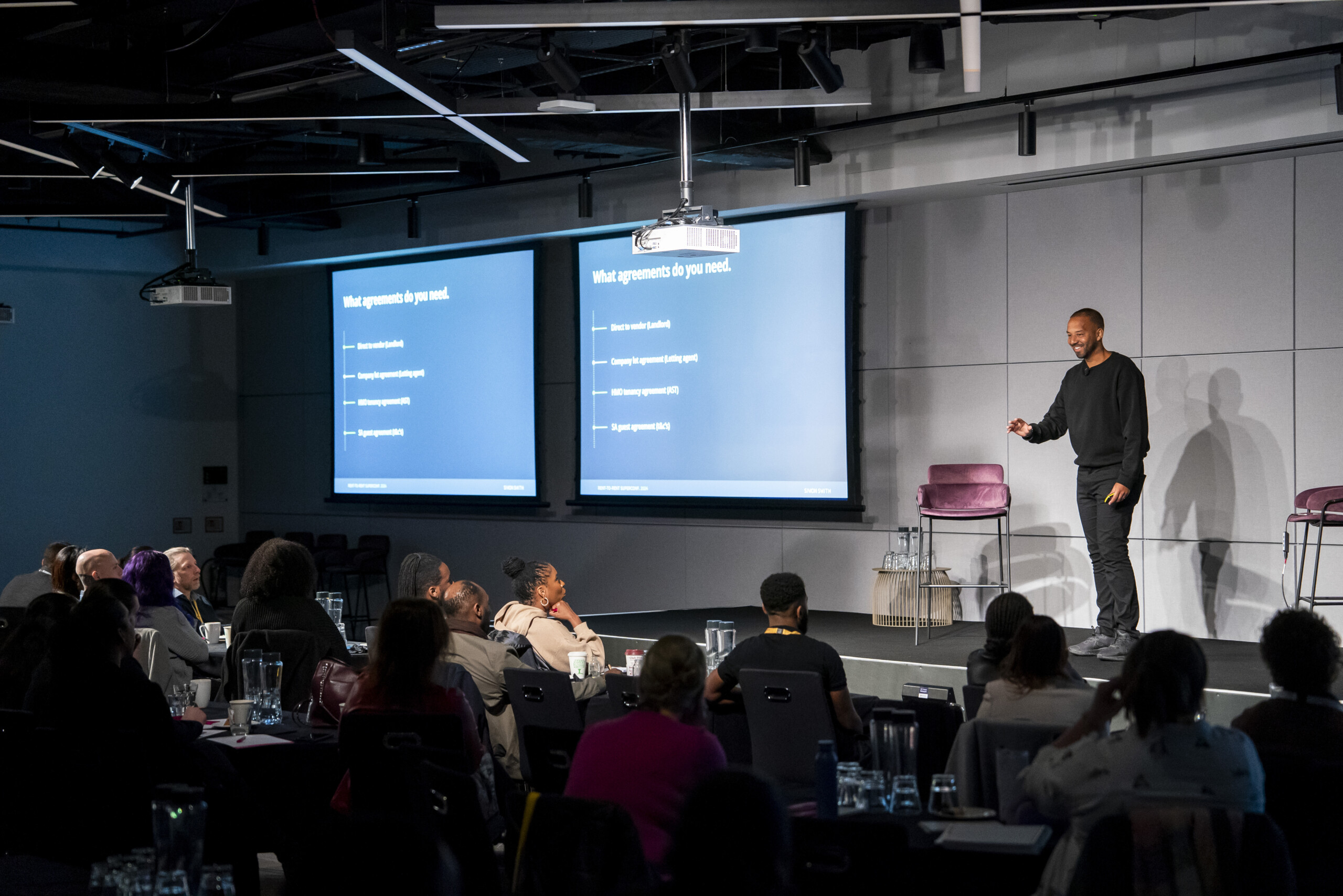 A speaker in a black outfit stands on a stage in front of an audience. Two large screens display a presentation titled "What agreements do you need." The audience is seated at tables with notebooks and glasses of water. James Gifford-Mead Photography - Event Photographer London