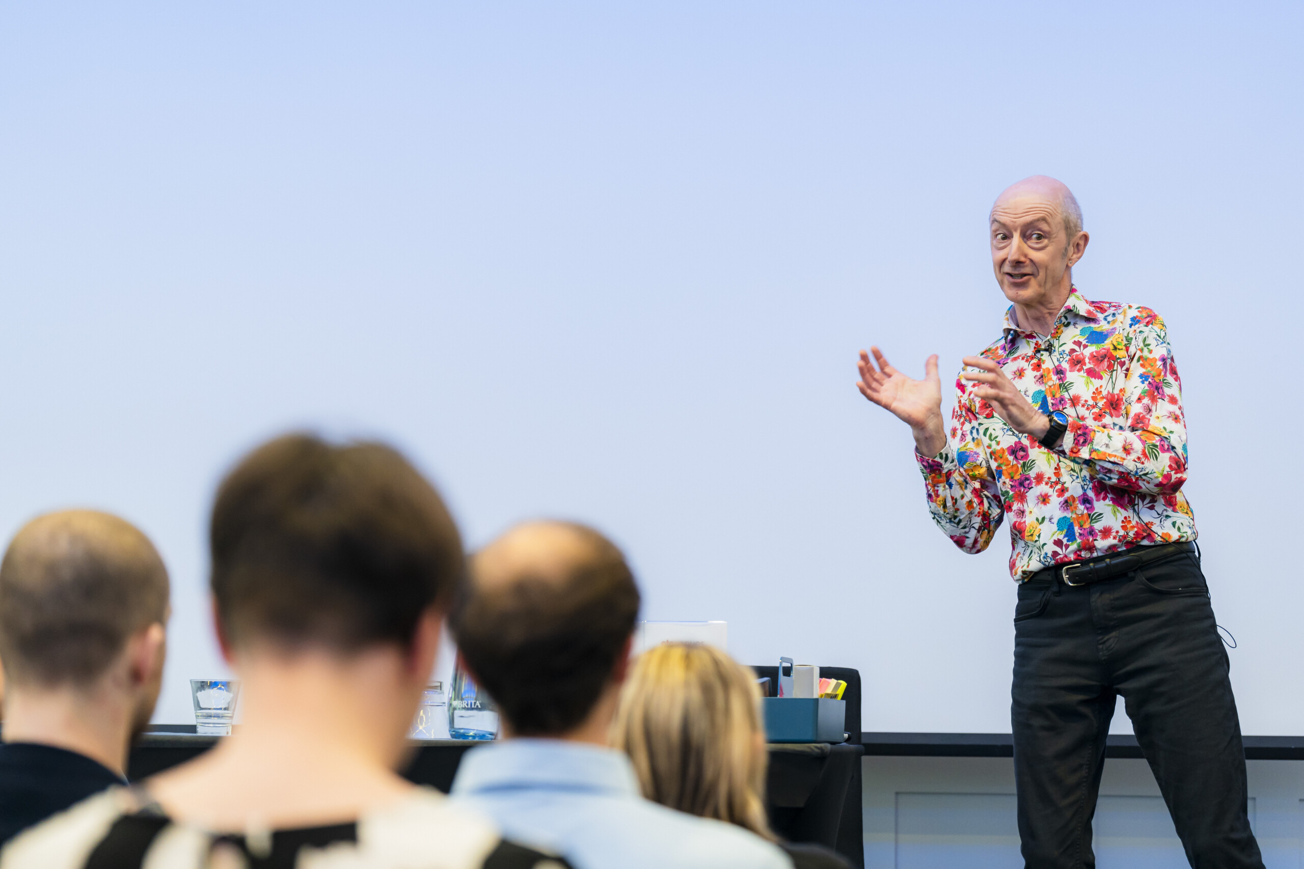 A man in a colorful shirt gestures enthusiastically while speaking to an audience. The audience's backs are visible in the foreground. He's standing in front of a white backdrop, possibly giving a presentation or lecture. James Gifford-Mead Photography - Event Photographer London