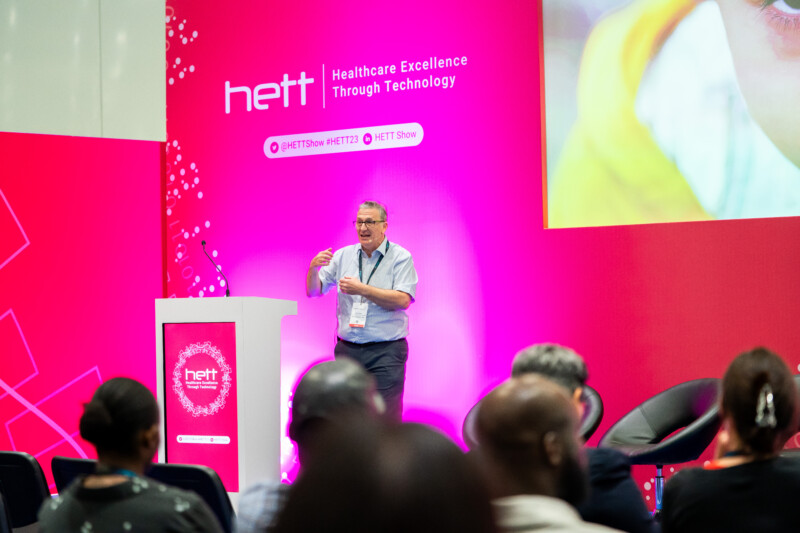 A speaker presents at a healthcare technology conference, captured brilliantly by a London event photographer. The stage, set against a pink backdrop with the "HETT" logo and tagline "Healthcare Excellence Through Technology," is abuzz as the audience eagerly listens from their seats. James Gifford-Mead Photography - Event Photographer London