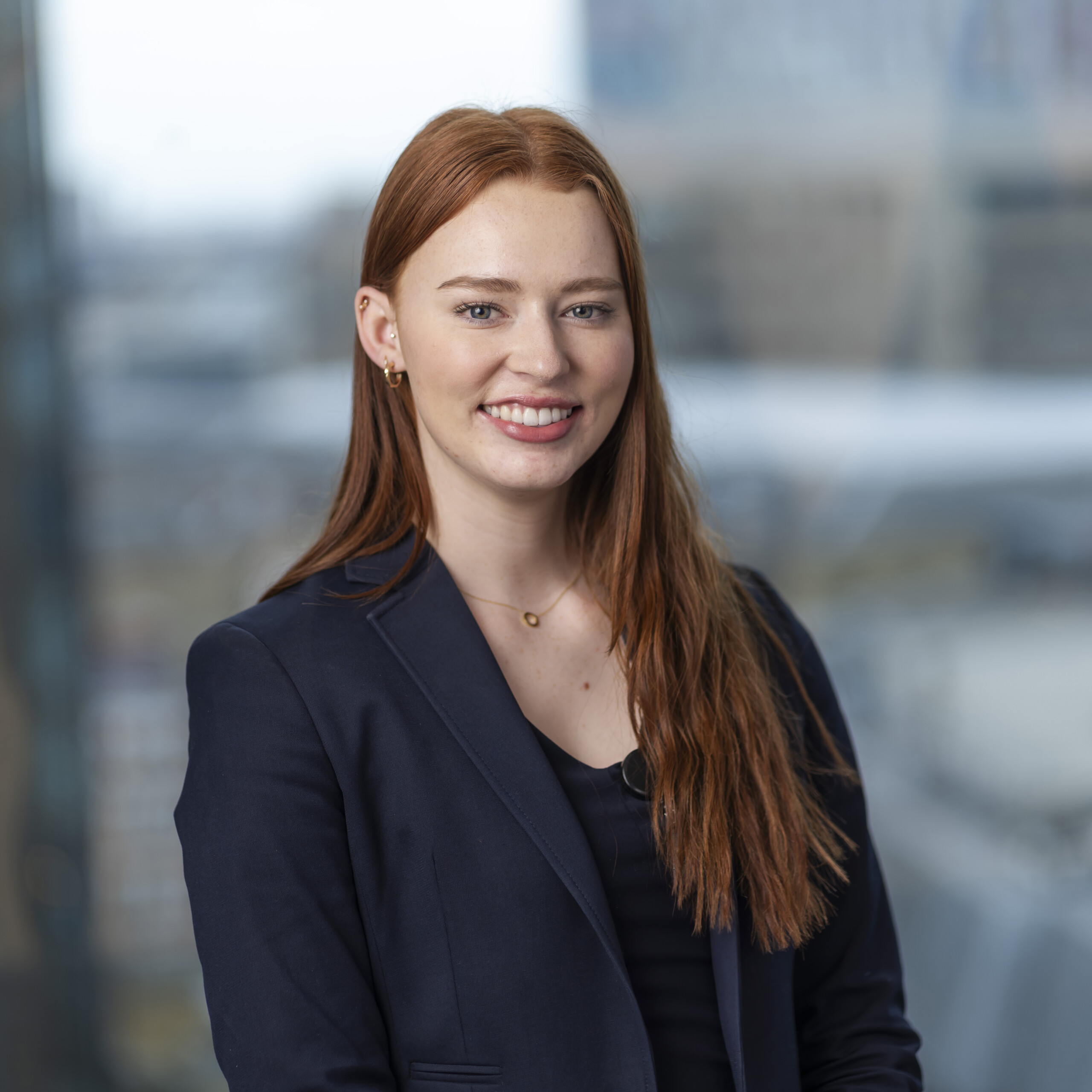 A person with long red hair is smiling, wearing a navy blazer over a black top. Captured by a corporate headshot photographer, they are indoors with a blurred cityscape visible through large windows in the background. James Gifford-Mead Photography - Event Photographer London