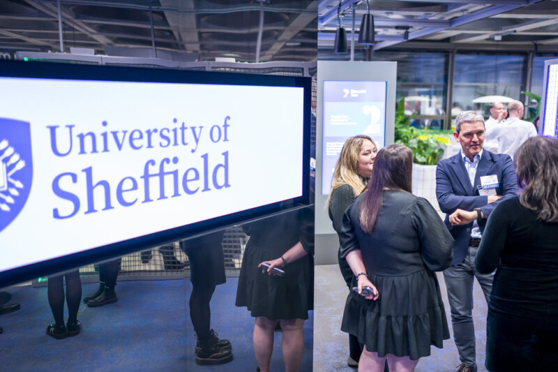 A group of people is engaged in conversation at an indoor event, captured by a London Conference Photographer. A large screen displays the text "University of Sheffield" on the left side. The setting appears modern and professional, with informational displays in the background. James Gifford-Mead Photography - Event Photographer London