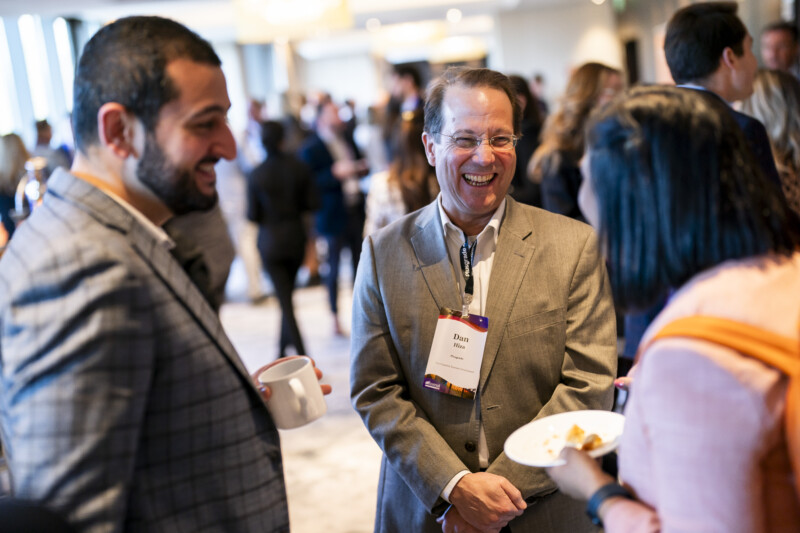 Three people in business attire are engaged in conversation at a networking event. A London Conference Photographer captures the man in the center smiling widely with a badge. The person on the left holds a coffee cup, while the one on the right balances a plate. A crowd bustles in the background. James Gifford-Mead Photography - Event Photographer London