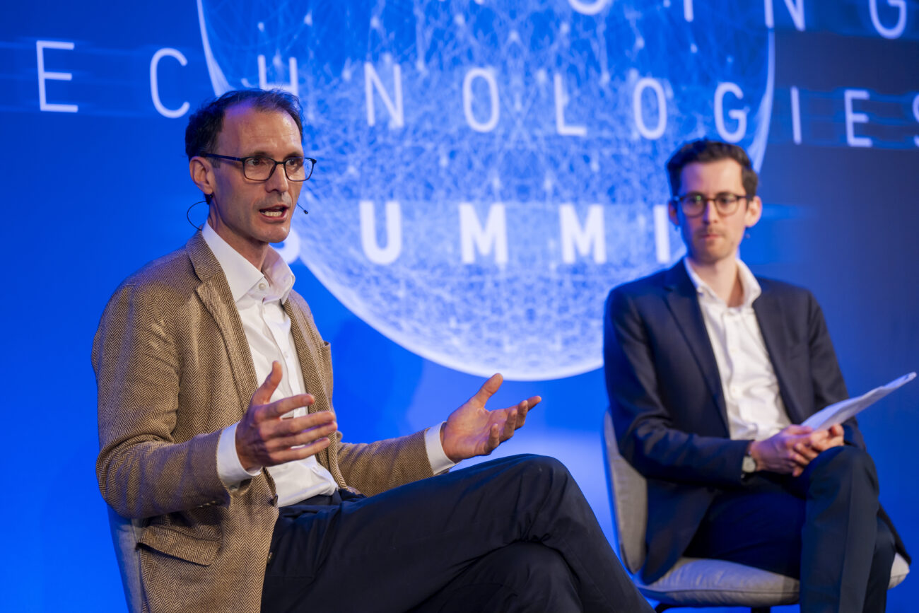 Two men in suits engage in discussion on stage at a London Technologies Summit. The man on the left gestures while speaking, and the man on the right holds a paper. A large, illuminated sign in the background reads "Technologies Summit," capturing a perfect moment for any conference photographer. James Gifford-Mead Photography - Event Photographer London