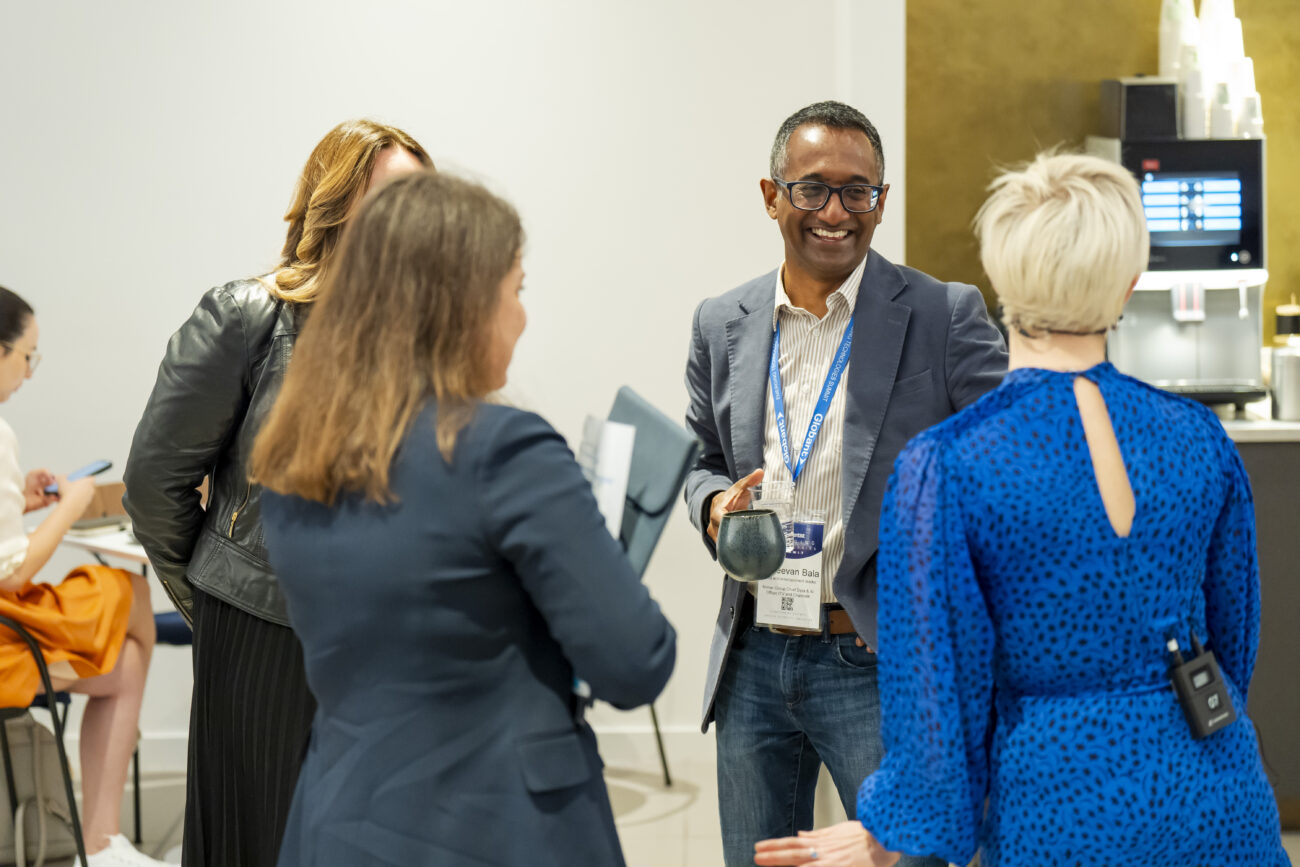 At a London conference, a photographer captures a lively office scene where a man with a lanyard and mug chats animatedly. The woman in the blue dress adds to the vibrant mix, while others casually converse, enhancing the lively atmosphere of collaboration. James Gifford-Mead Photography - Event Photographer London