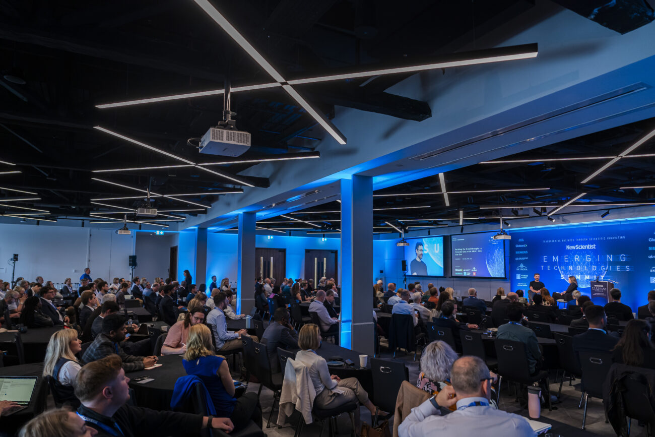 A large conference room brims with seated attendees, attentively listening to a panel discussion. Bathed in blue lighting, the room showcases presentations on vibrant screens. Captured by a London Conference Photographer, the engaged audience faces the stage where dynamic speakers hold sway. James Gifford-Mead Photography - Event Photographer London