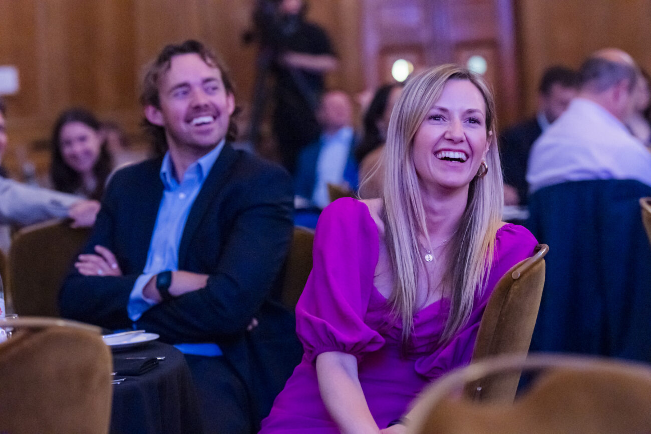 At a lively gathering, a woman in a purple dress and a man in a suit sit together, smiling and laughing. Captured by an event photographer, the scene showcases the vibrant essence of London corporate photography