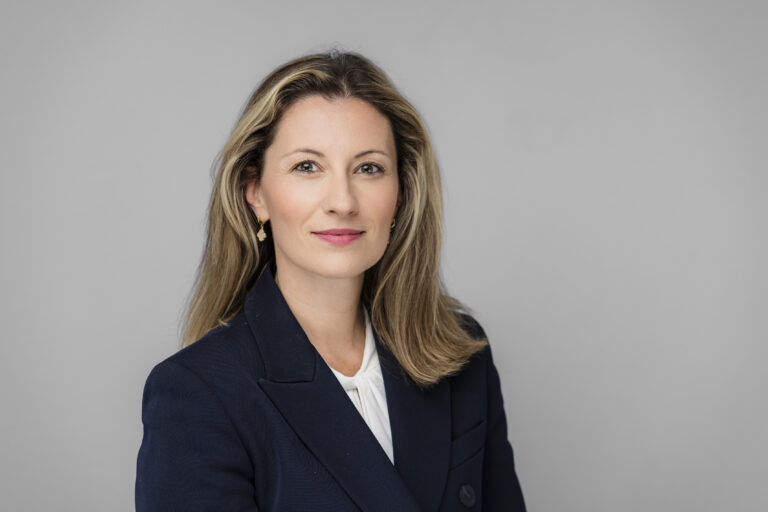 A woman with long, light brown hair and wearing a dark blazer over a white blouse poses for a corporate headshot. She is set against a plain gray background, maintaining a calm expression as she faces forward, expertly captured by the photographer