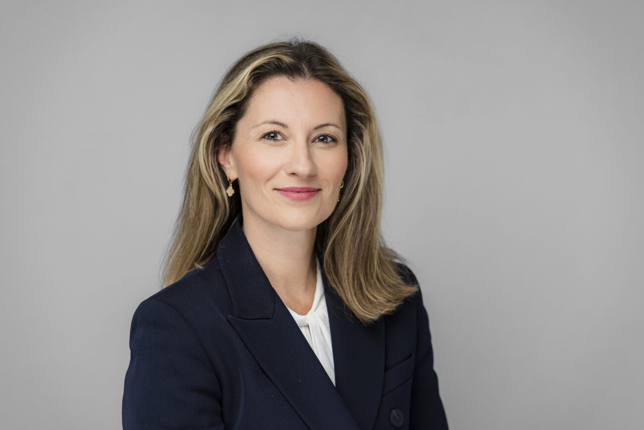 A woman with long blonde hair, captured by a skilled corporate headshot photographer, wears a dark blazer and light shirt, smiling softly at the camera against a plain gray background. James Gifford-Mead Photography - Event Photographer London