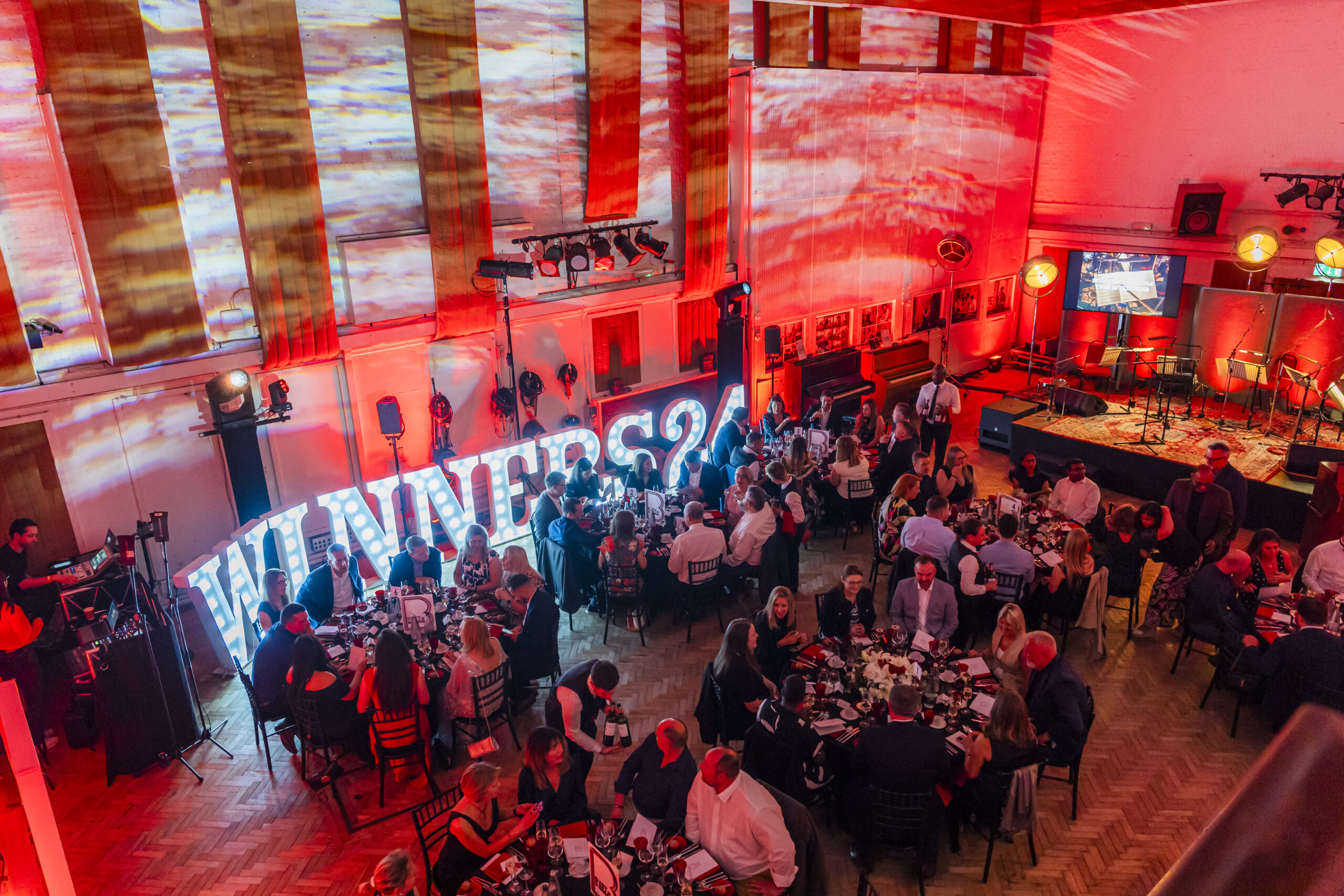 A red and warmly lit banquet hall features crowded tables with guests. Large illuminated letters spell "WINNERS" in the background. A stage with musical instruments is set up on the right. Many attendees are engaged in conversation. James Gifford-Mead Photography - Event Photographer London