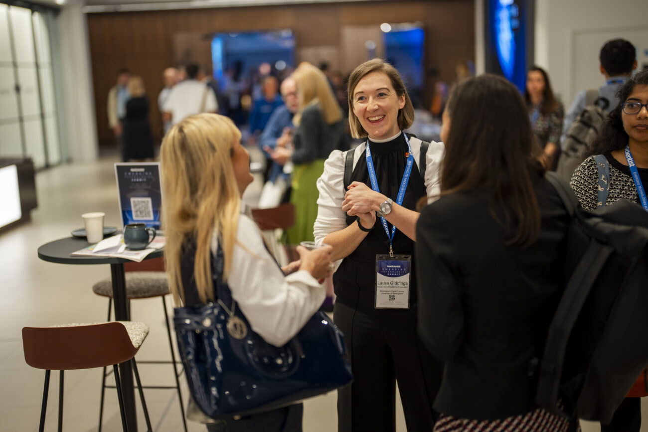A group of people is engaged in conversation at a networking event. They are standing indoors, wearing business attire and event badges. A table with a coffee cup and a brochure is nearby. The atmosphere is lively and professional. James Gifford-Mead Photography - Event Photographer London