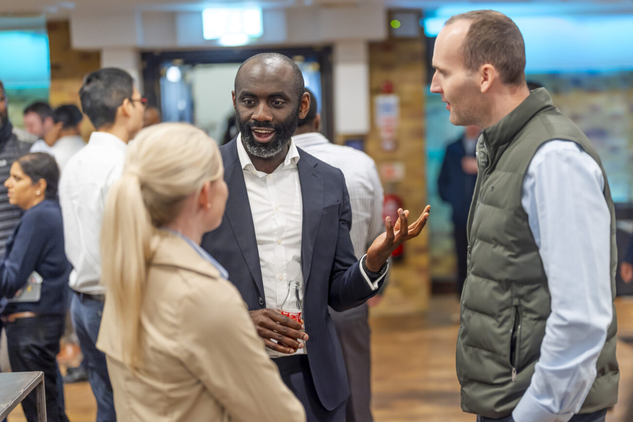 A group of people engaging in conversation at a social event. A man in a suit gestures animatedly while holding a drink, accompanied by a woman in a beige coat and a man in a vest with folded arms. Other attendees are visible in the background. James Gifford-Mead Photography - Event Photographer London