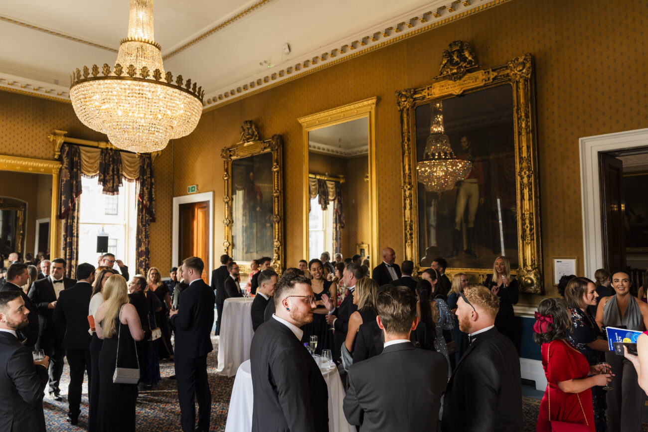 A formal gathering in an elegant room with chandeliers, large mirrors, and ornate gold-framed paintings. Attendees in formal attire mingle and converse around tall tables with white tablecloths. James Gifford-Mead Photography - Event Photographer London