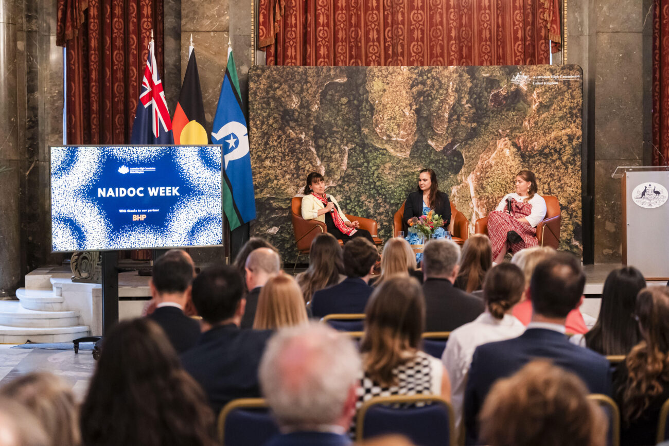Three people sit on stage in discussion chairs, with a screen displaying "NAIDOC Week" beside them. Flags and a large map serve as the backdrop. Audience members are seated in the foreground. James Gifford-Mead Photography - Event Photographer London