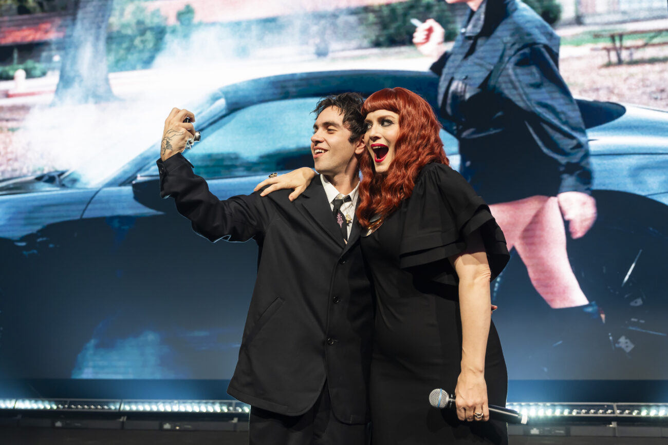 A man and a woman with red hair are taking a selfie on stage, smiling. They are dressed in formal black attire, standing in front of a large screen displaying a scene with a car and a person in the background. James Gifford-Mead Photography - Event Photographer London