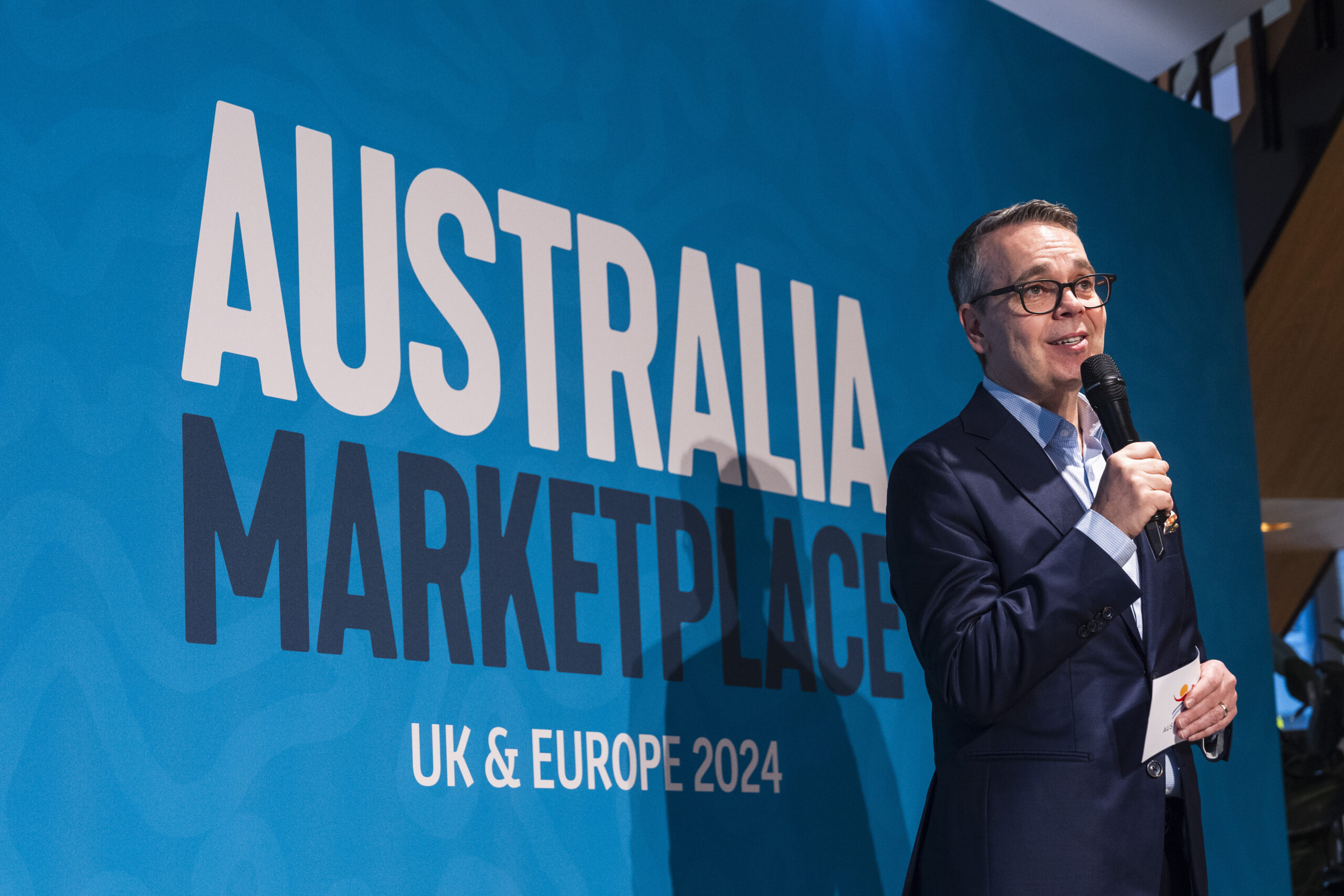 A person in a suit speaks into a microphone next to a large blue sign that reads "AUSTRALIA MARKETPLACE UK & EUROPE 2024. James Gifford-Mead Photography - Event Photographer London