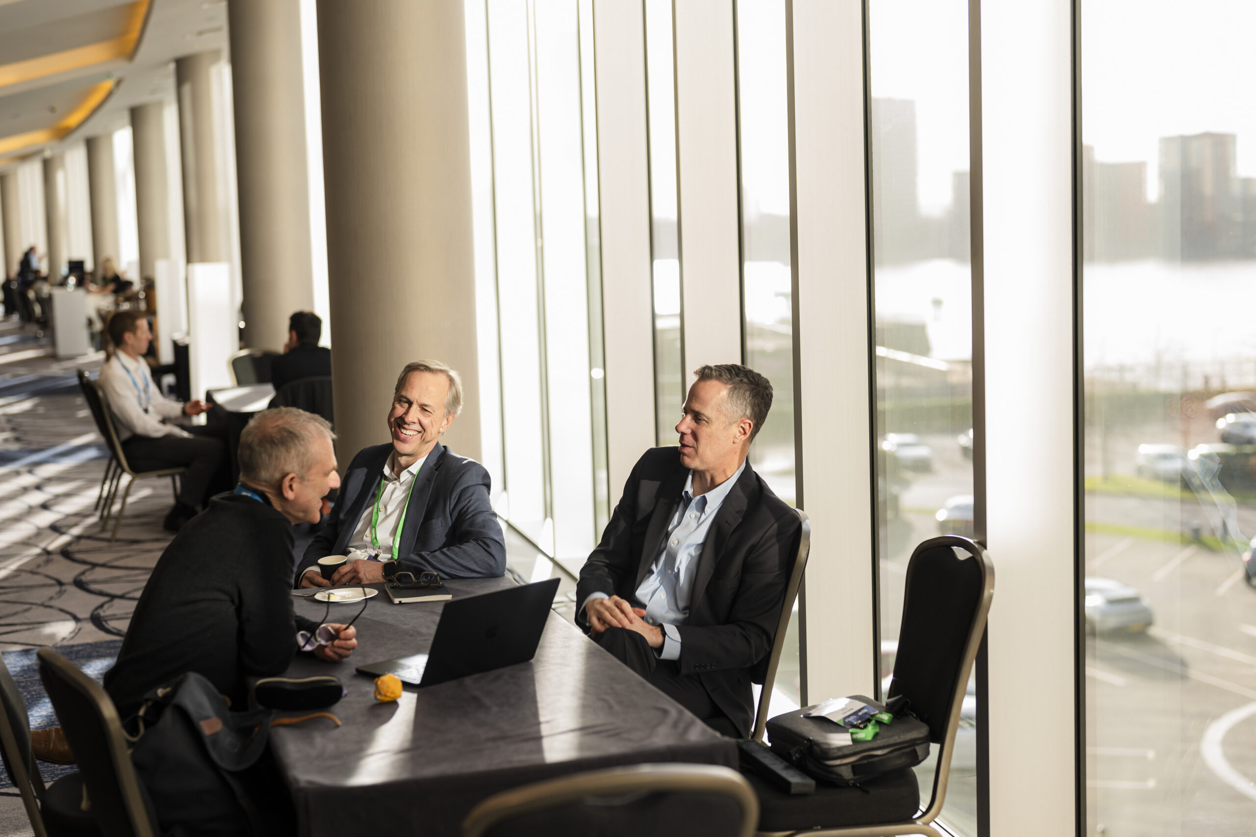 Three men in business attire sit at a table with laptops and papers in a sunlit conference hallway. Large windows reveal a cityscape. The setting appears professional, with others in the background having discussions. James Gifford-Mead Photography - Event Photographer London