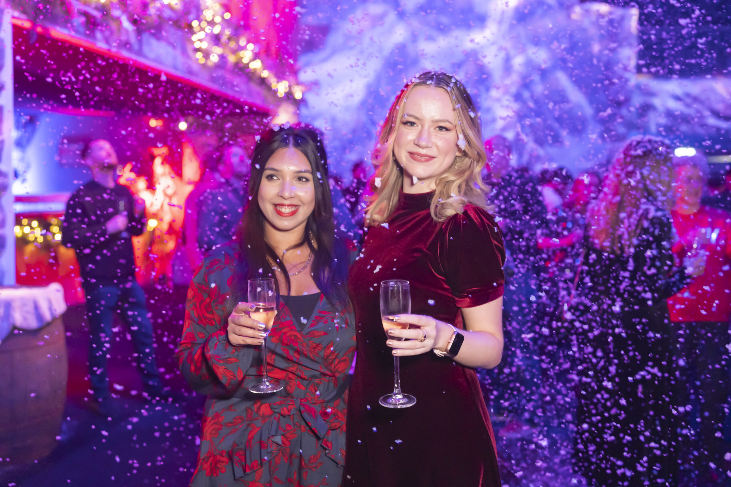 Two women in festive attire hold champagne glasses at a holiday-themed event. Snowflakes fall around them. The background is filled with people, lights, and holiday decorations, creating a joyful atmosphere. James Gifford-Mead Photography - Event Photographer London