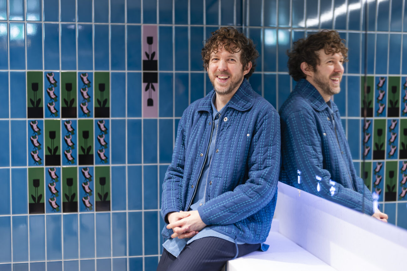 Man smiling seated against patterned tiled wall.