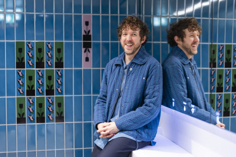 Man smiling seated against patterned tiled wall.