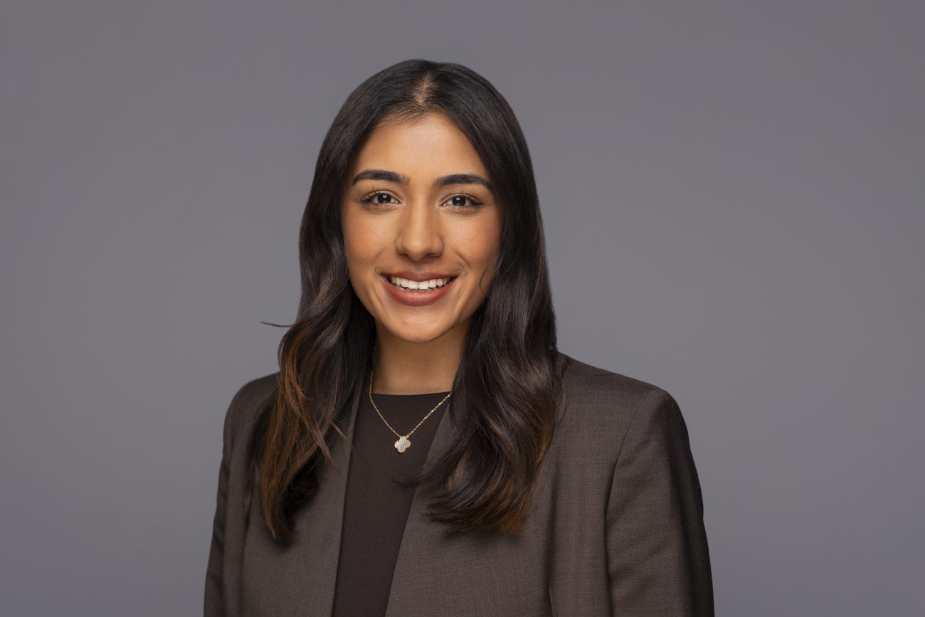 A woman with long dark hair, wearing a dark blazer and a gold necklace, smiles in front of a plain grey background—an ideal example of the polished results you can expect from a professional corporate headshot photographer