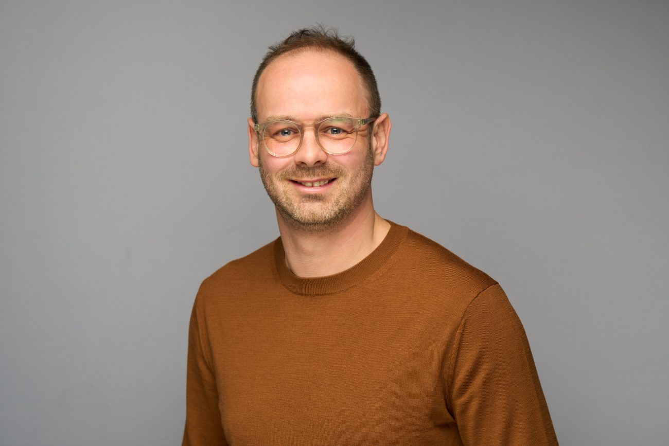 A smiling man with short hair, glasses, and a beard, wearing a brown jumper, stands in front of a plain grey background—an ideal example of work by a skilled corporate headshot photographer