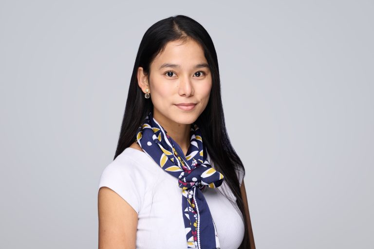 A woman with long black hair wears a white top and a blue patterned scarf, standing against a plain light grey background. Captured by a Corporate Headshot Photographer, she faces the camera with a slight smile